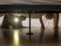 Mother and daughter, babysitter and kid girl afraid of monsters hold smartphone shine a flashlight under the bed looking on floor check ghosts, older younger sisters playing having fun at home concept