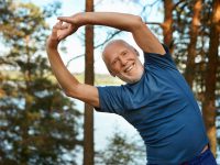 Outdoor shot of happy energetic senior retired man enjoying physical training in park, doing side bends exercise, holding hands together, looking at camera with broad smile, warming up body before run